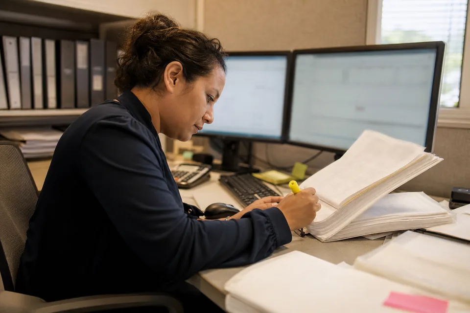Credit union accounting analyst comparing a printed core banking report with general ledger balances on two monitors
